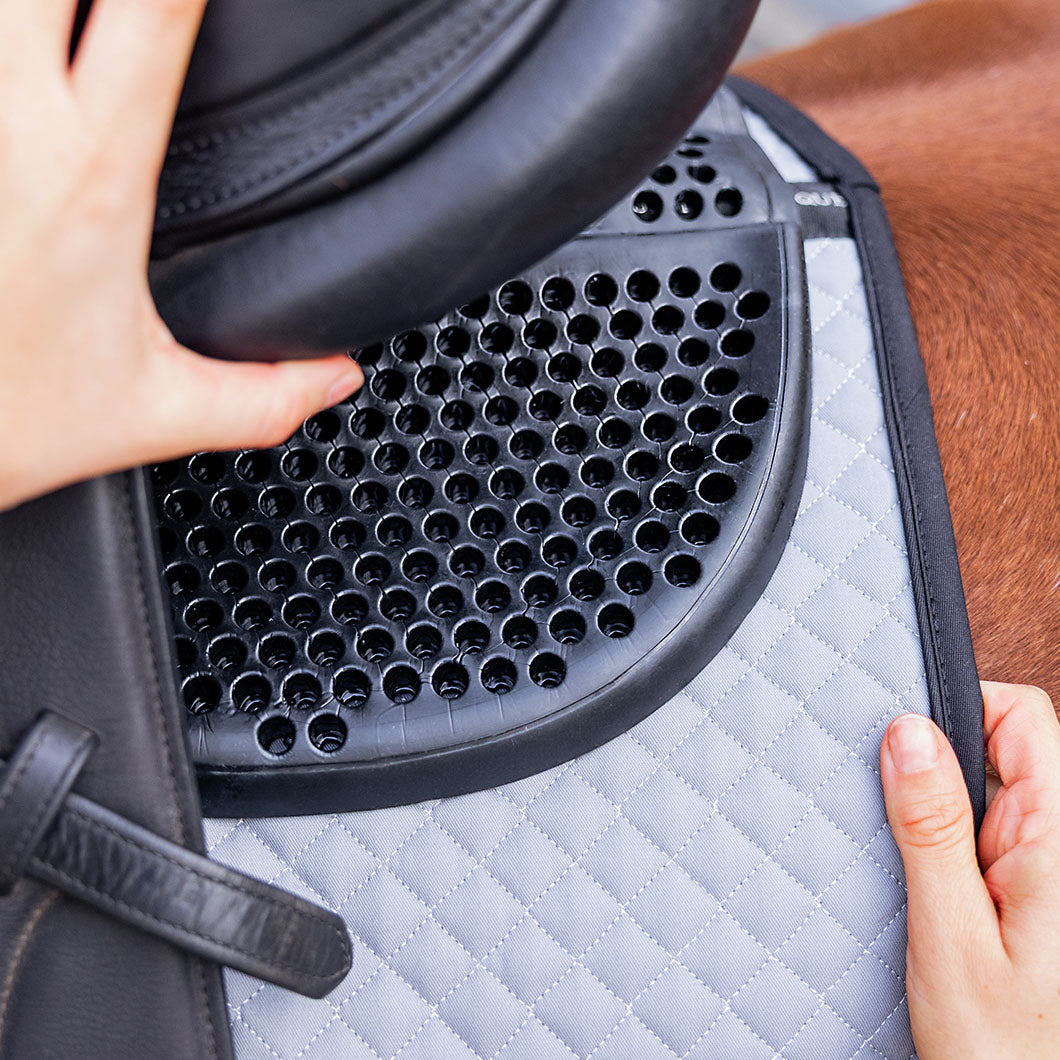 Hand hebt den Sattel an und zeigt das EQUEST Gel Pad Comfort mit Lochstruktur auf einer grauen Schabracke